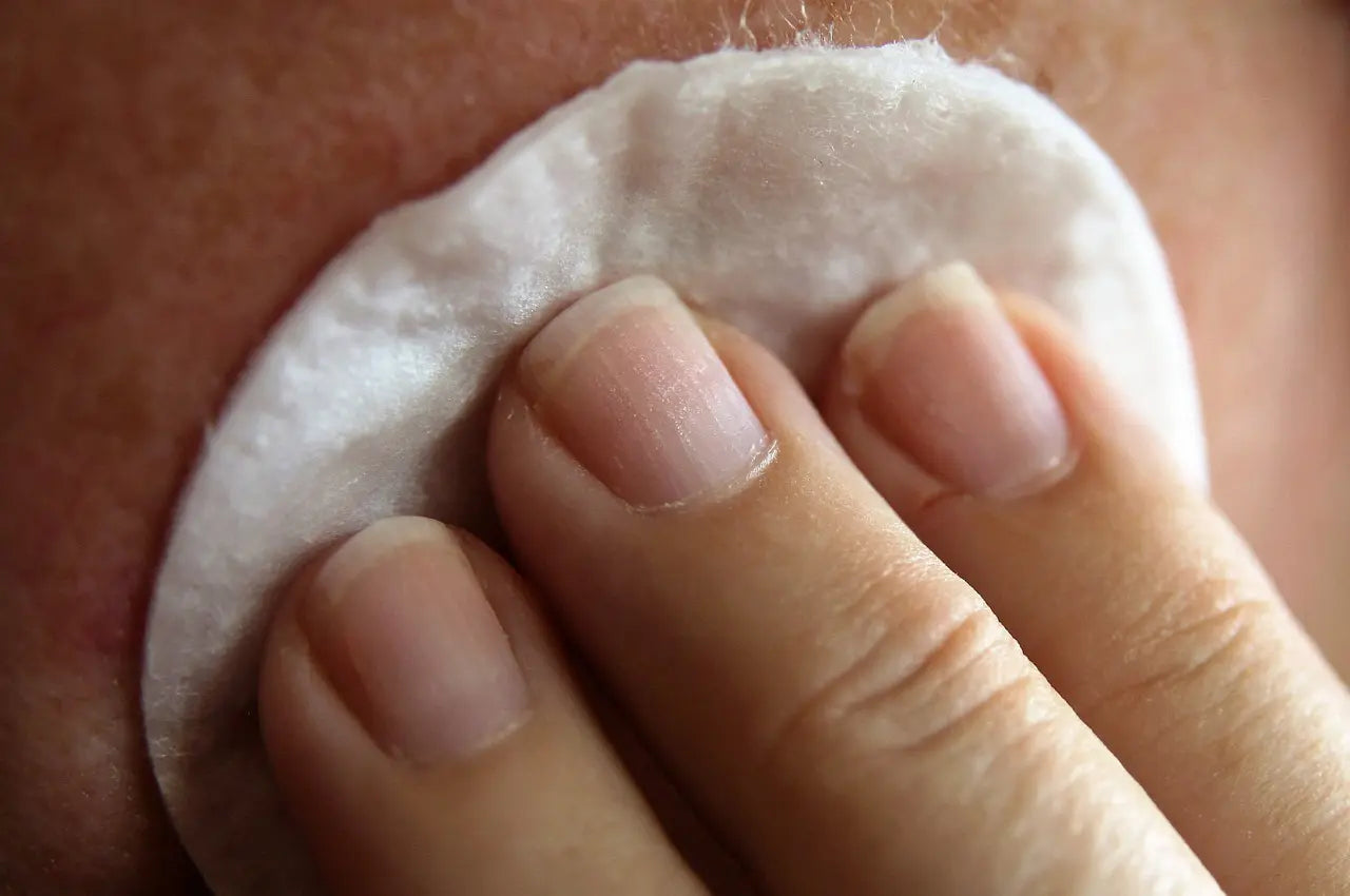 Primer plano de una mano sosteniendo un disco de algodón mientras limpia la piel del rostro. La imagen muestra detalles de la piel y las uñas naturales de la persona.