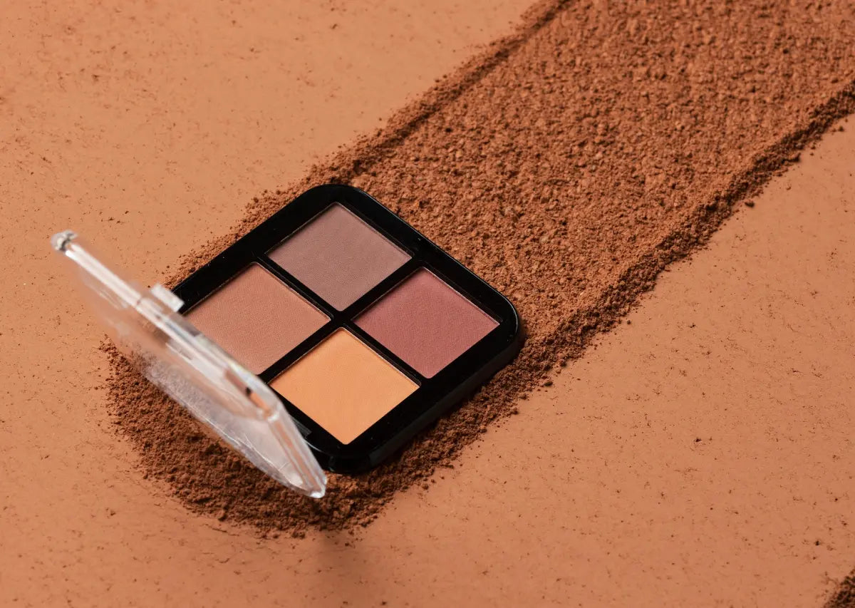 Paleta de sombras de ojos en tonos tierra sobre un fondo de polvo marrón. La paleta contiene cuatro colores mate en una caja negra con tapa transparente.