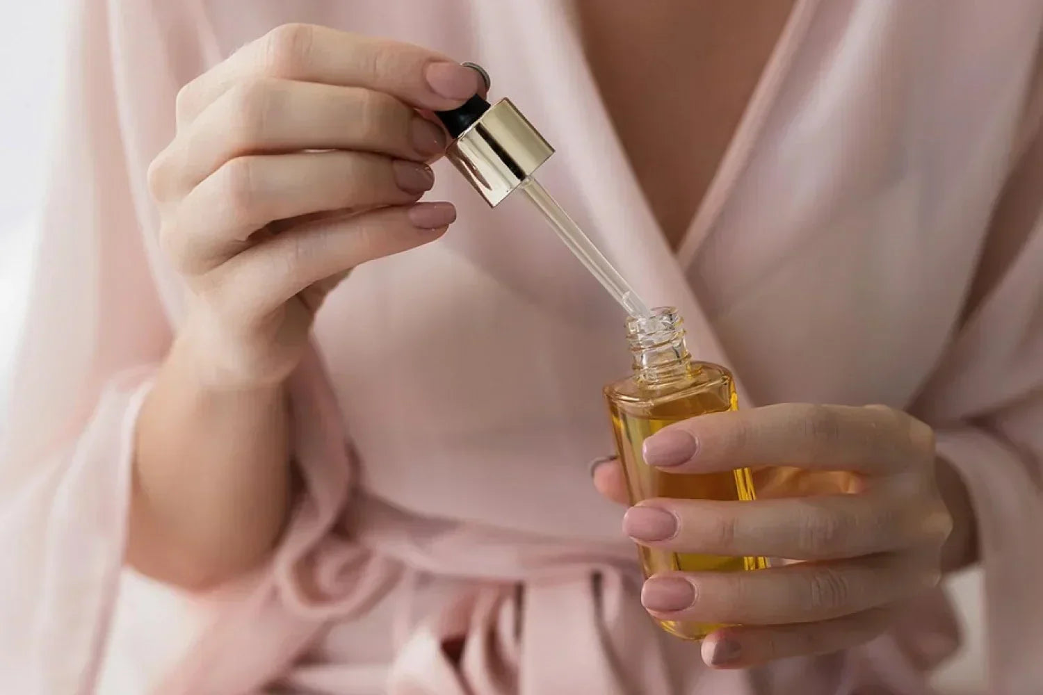 Manos femeninas sosteniendo un frasco de vidrio con sérum de color ámbar y aplicador de gotero dorado, mientras una mujer viste una bata rosada.
