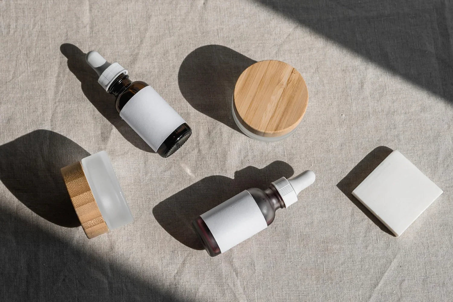 Frascos de vidrio con goteros y tarros de crema con tapas de bambú sobre tela beige, ideales para cosmética natural y ecológica.